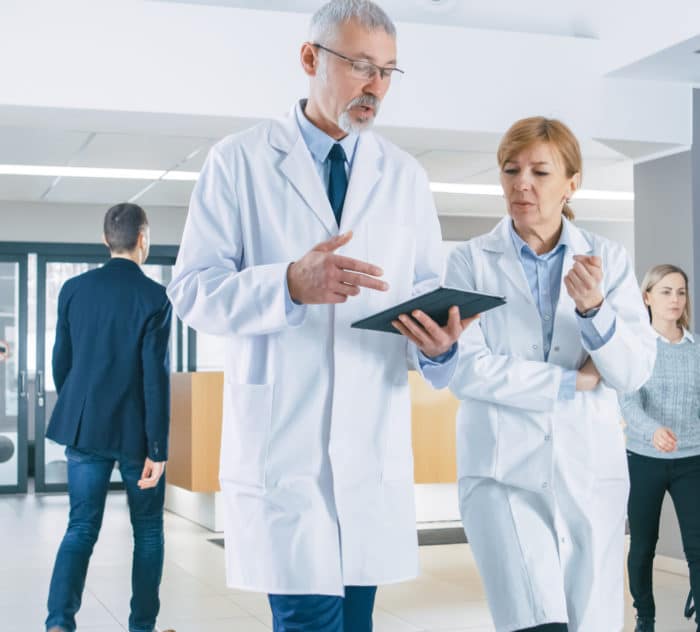 In the Hospital, Busy Doctors Talk, Using Tablet Computer While Walking Through the Building. New Modern Fully Functional Medical Facility.